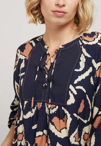 Blouse marine avec des motifs abstraits beiges et rouille, dotée d'un col rond, de détails à nouer et de tissu froncé pour un ajustement ample.