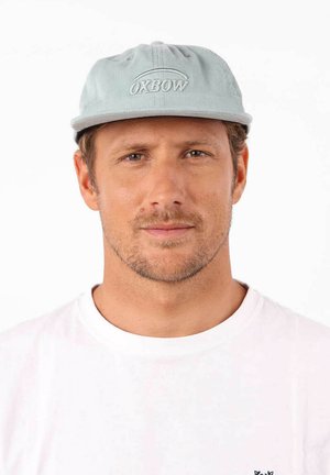 Casquette en velours côtelé bleu clair avec le logo brodé "OXBOW" à l'avant. Texture douce, forme arrondie et visière plate. Portée avec un t-shirt blanc.