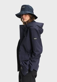 Femme portant une veste à capuche bleu marine et un bob assorti, tous deux avec des écussons visibles de la marque Aigle, debout devant un fond clair.