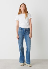 Uitlopende blauwe jeans met een middelmatig gewassen denimtextuur, voorzien van vijf zakken en een knoopsluiting. Gekleed met een eenvoudig wit T-shirt.