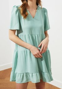 Mintově zelené šaty s krátkými balónovými rukávy, výstřihem do V a vrstveným volánovým lemováním. Vyrobené z lehké tkaniny s jemnou texturou.