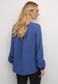 Blauwe blouse met lange mouwen en een lichte glans, met een ronde zoom, geplooide details aan de achterkant en elastische manchetten. Geverfde stof.