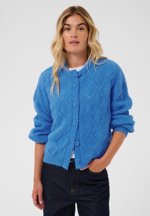 Femme aux longs cheveux blonds portant un cardigan bleu boutonné avec un motif en losange et un jean foncé, debout avec une main dans la poche.