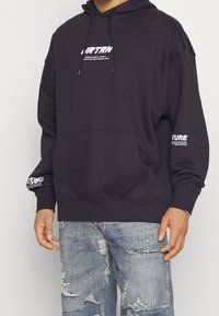 Hombre vestido con una sudadera negra con texto blanco en el pecho y las mangas, combinada con unos jeans rotos de color azul claro.