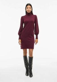 Burgunderfarbendes Turtleneck-Kleid mit langen, gepufften Ärmeln. Glatte Strickstruktur, oberhalb des Knies, kombiniert mit passgenauen schwarzen Stiefeln bis zum Knie.