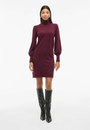 Vestido burdeos de cuello alto con mangas largas y abullonadas. Textura de punto suave, con longitud por encima de la rodilla, combinado con botas negras ajustadas hasta la rodilla.