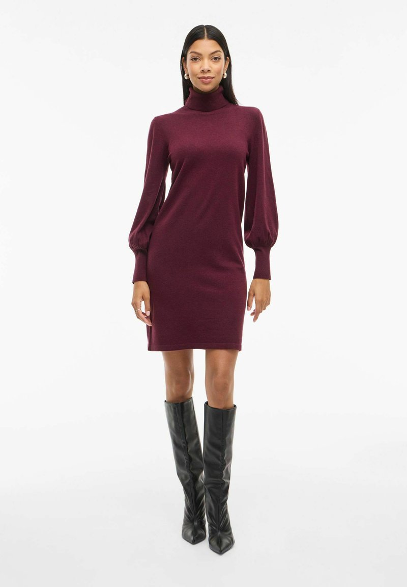 Burgunderfarbendes Turtleneck-Kleid mit langen, gepufften Ärmeln. Glatte Strickstruktur, oberhalb des Knies, kombiniert mit passgenauen schwarzen Stiefeln bis zum Knie.