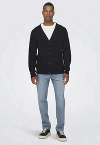 Cardigã preto canelado com decote em V, apresentando cinco botões na frente, combinado com jeans azul claro e sapatilhas pretas.