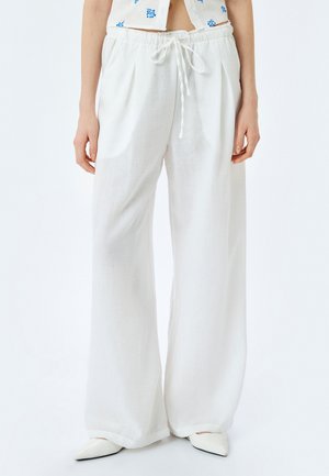 Pantalons larges en lin blanc avec taille à cordon, présentant une texture douce et une coupe décontractée. Associés à des chaussures blanches à bouts pointus.