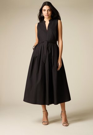 Vrouw draagt een mouwloze zwarte midi-jurk met tailleband en patterned hoge hakken, staand tegen een beige achtergrond.