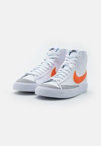 Ett par vita hög-top Nike-sneakers med orange swoosh-logotyper, grå tåskydd, vita snören och vita texturerade sulor på en vit bakgrund.