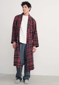 Karirani bathrobe u crvenoj, plavoj i bijeloj boji, dugih rukava, do koljena. Uparen s plavim i bijelim prugastim izborima hlača za spavanje i sivim papučama.