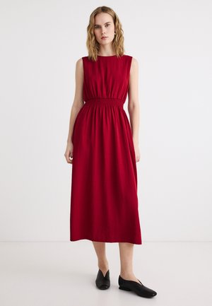 Woman standing wearing sleeveless red midi dress with gathered waist and black flat shoes against plain white background.