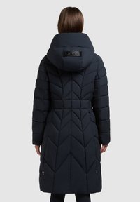 Vrouw die een lange, doorgestikte zwarte donsjas draagt met een grote capuchon en een zichtbaar merkembleem op de achterkant, staand tegen een effen achtergrond.