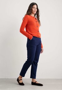 Oranžový vlněný svetr s kulatým výstřihem a pěti knoflíky, kombinovaný s tmavě modrými slim-fit kalhotami. Modelka má na sobě tmavě hnědé ploché boty.