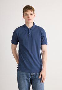 Joven vestido con una camiseta polo azul marino y jeans azules, de pie contra un fondo claro liso, mirando hacia adelante con una expresión neutral.