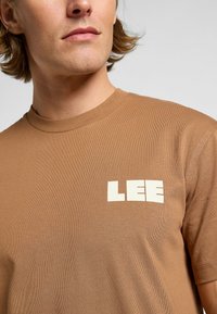 Brun bomull t-shirt med rund hals och en iögonfallande vit "LEE"-logotyp på vänster bröst, som visar en ribbad struktur.
