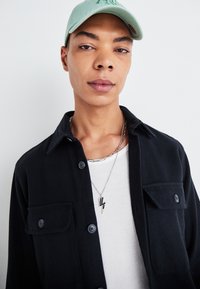 Chemise noire à boutons avec deux poches avant sur une chemise blanche à côtes. Collier en forme d'éclair argenté et casquette vert menthe. Texture de peau lisse.