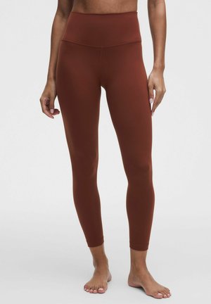 Femme portant un legging marron taille haute, debout pieds nus devant un fond clair uni.