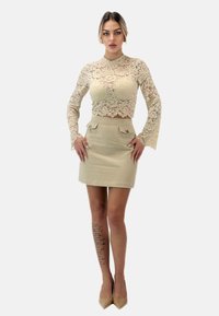 Top corto in pizzo beige con orlo scallopato e maniche lunghe, abbinato a una gonna beige alta in vita con due tasche frontali e dettagli con bottoni.