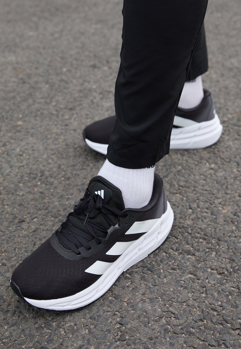 Chaussures de sport noires avec des accents blancs, tige en mesh et semelle texturée. Portées avec des chaussettes blanches et un pantalon de sport noir sur une surface texturée.