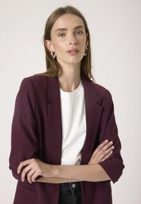 Burgundi bleiser, mille struktureeritud õlgade ja volditud varrukatega, kantud valge särke peal. Mudel poseerib käed risti, neutraalne taust.