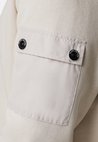 Felpa beige chiaro caratterizzata da un corpo testurizzato e maniche in tessuto liscio, con tasca a patta, bottone a pressione nero e dettagli cuciti.