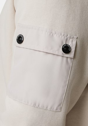 Felpa beige chiaro caratterizzata da un corpo testurizzato e maniche in tessuto liscio, con tasca a patta, bottone a pressione nero e dettagli cuciti.