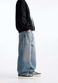 Zwarte bomberjack met geribbelde manchetten, gedragen over een wit shirt. Losvallende lichtblauwe jeans en zwarte sneakers maken de outfit compleet.