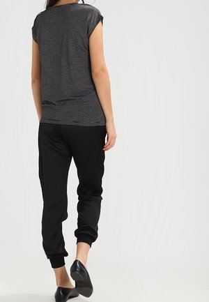 Femme portant un haut à manches courtes noir et blanc rayé, un pantalon de jogging noir et des ballerines noires, debout de dos sur un fond uni.