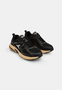 Scarpe sportive nere con dettagli in rete, suole in gomma e accenti beige. Presentano un design con lacci e una linguetta posteriore per facilitare l'indossamento.