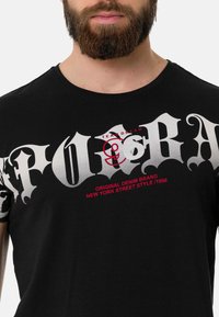 Schwarzes Baumwoll-T-Shirt mit kräftigen weißen Lettern und roten Akzenten. Das Design enthält den Markennamen, die Modellnummer und ein grafisches Element.