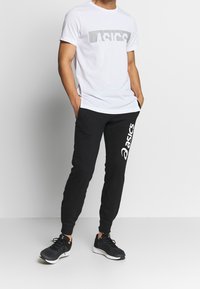 Bílá tričko s šedým logem ASICS, doplněné černými tepláky s bílým logem ASICS. Černé sportovní boty se šněrováním.