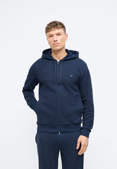 Mornarska zip-up majica izrađena od mekanog materijala, s kapuljačom na vezanje, rebrastim manžetama i rubom, te malim logotipom na prsima.