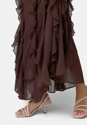 Falda midi marrón con volantes combinada con sandalias de tacón alto rosa claro con tiras, en una persona de pie.