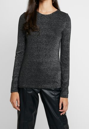 Kvinde iført en tætsiddende langærmet sølvglitrende top med sorte læderbukser, stående mod en ensartet lys baggrund.