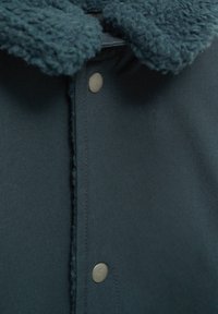 Mörkt tealfärgad tygjacka med mjuk fleecekrage och två synliga silverknappar vid framstängningen.