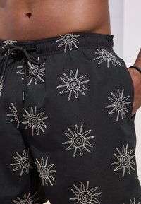 Pantaloncini da bagno neri con ricamo bianco a forma di sole, vita elastica con coulisse, tasche laterali e tessuto dalla texture liscia.