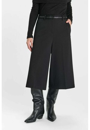 Frau trägt schwarze Culottes mit weitem Bein und schwarzem Gürtel, schwarzes langärmeliges Oberteil und schwarze kniehohe Lederstiefel, steht mit Händen in den Taschen.