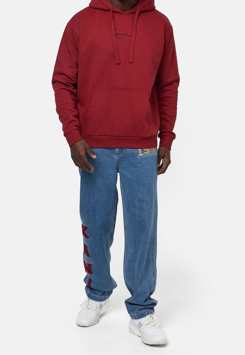 Roter Hoodie mit einer Fronttasche und einem schwarzen Logo; kombiniert mit blauen Denim-Hosen mit seitlichem Print und lockerem Schnitt. Weiße Sneakers.