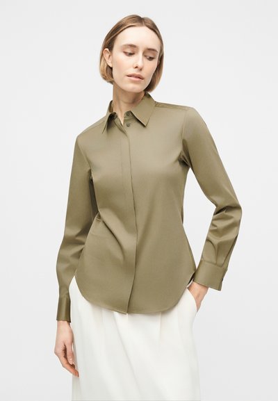 Γυναίκα που φοράει εφαρμοστό πουκάμισο μακριά μανίκια σε ελιά πράσινο χρώμα με άσπρο παντελόνι, στέκεται μπροστά σε απλό ανοιχτόχρωμο φόντο.
