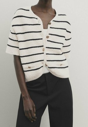 Personne portant un cardigan blanc à manches courtes avec des rayures horizontales noires et des boutons, associé à un pantalon noir, sur un fond uni.