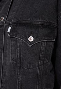 Tascone di giacca in denim nero con un bottone argento inciso con il testo del marchio e un piccolo etichetta di tessuto sul lato sinistro della cucitura.