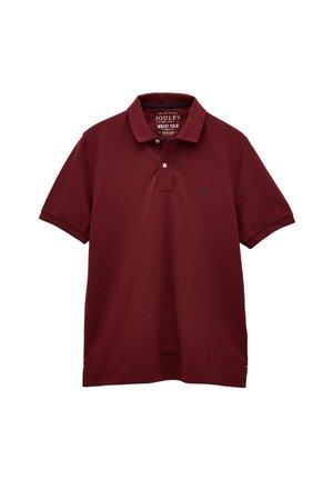 Mand iført en bordeaux polo, beige bukser og brune sko, der står op ad en beige tekstureret væg med den ene hånd i lommen.