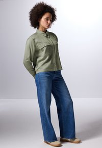 Camicia verde chiaro a maniche lunghe con tasche sul petto; abbinata a jeans a gamba larga blu; la modella sta in piedi con le mani sui fianchi contro uno sfondo neutro.