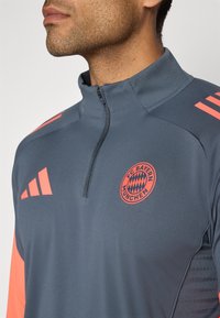 Sivi atletski pulover s patentnim zatvaračem, s narančastim prugama i logotipom FC Bayern München. Izrađen od glatkog, laganog materijala.