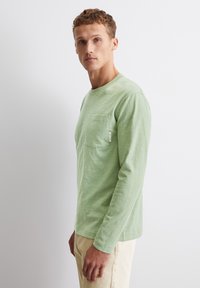 Hellgrünes Langarm-Shirt mit rundem Ausschnitt, das eine Brusttasche und eine subtile Textur aufweist. Kombiniert mit beigen Hosen.