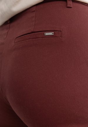 Burgundi värvi püksid, mis on valmistatud siledast kangast, millel on tagakotikeses hõbedane metalne silt. Disain on sobiv ja puhta viimistlusega.