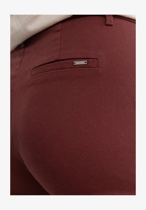 Burgundi värvi püksid, mis on valmistatud siledast kangast, millel on tagakotikeses hõbedane metalne silt. Disain on sobiv ja puhta viimistlusega.