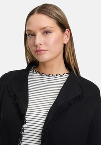 Černý svetr s třásněmi, nošený přes černobílý pruhovaný top. Hladká textura, design s otevřeným předním dílem, dlouhé rukávy.